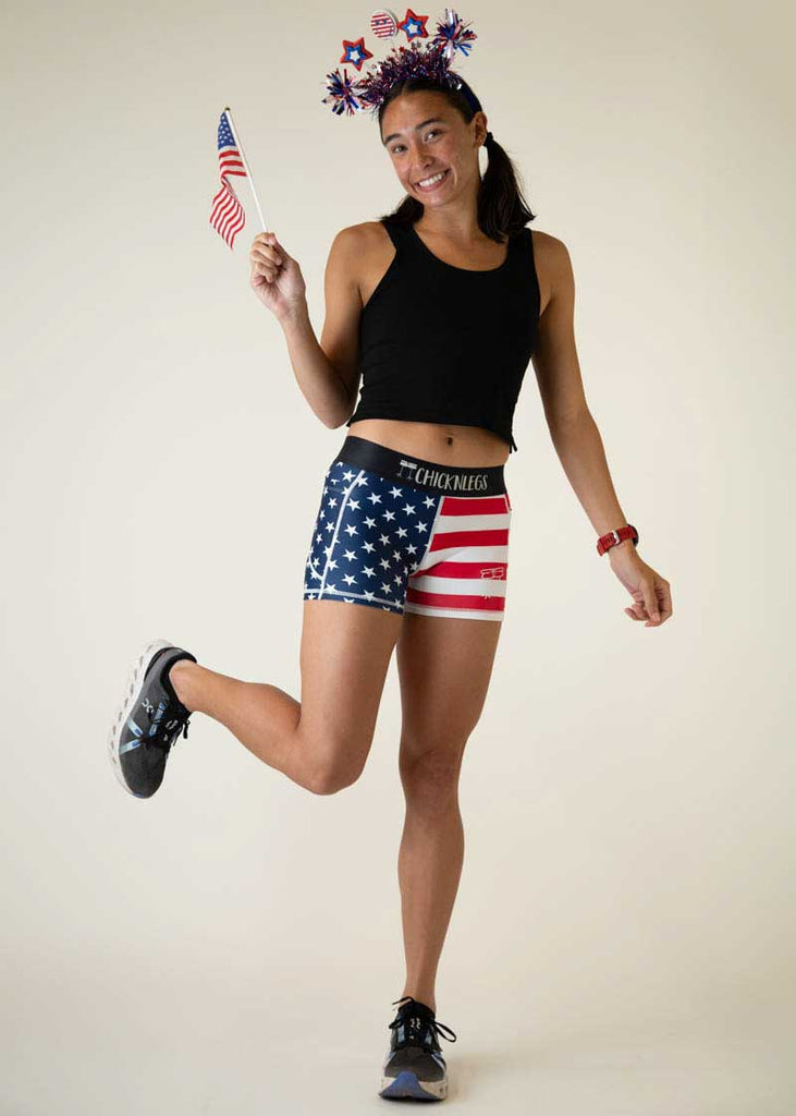 Full body view of the women's 3 inch USA compression running shorts from ChicknLegs holding a mini flag and lifting one leg up.