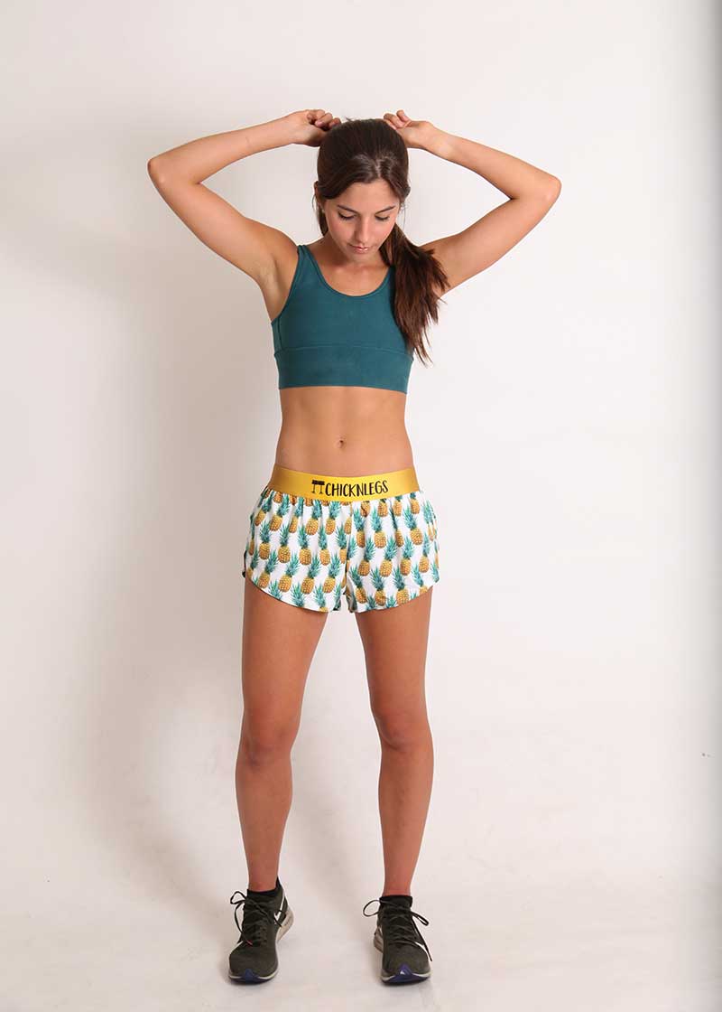 ChicknLegs women's trippy pineapples 1.5" split running shorts full body shot with green sports bra and fixing hair.